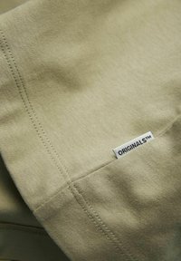 Tejido verde oliva con una textura suave, que presenta un detalle de costura y una pequeña etiqueta "ORIGINALS™" cosida en el borde.