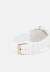 Correa de reloj de silicona blanca con hebilla de metal en tono dorado. La parte trasera presenta una superficie circular lisa con un patrón texturizado.