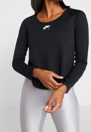 Kobieta ubrana w czarną, długorękawową bluzę Nike Air i jasnoszare legginsy Nike, poprawiająca rękaw, na jednolitym jasnym tle.