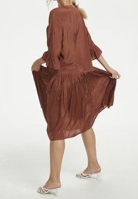 Brun, flydende kjole lavet af let stof med lange ærmer med flæseelementer og en samlet talje, knælængde.