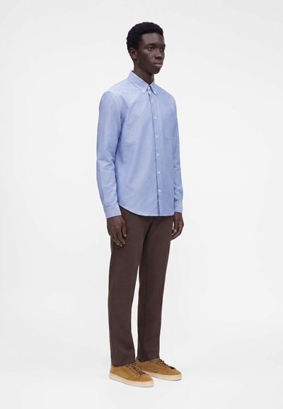 Homme debout portant une chemise bleu clair à manches longues boutonnée, un pantalon marron foncé et des baskets en daim beige sur fond blanc.