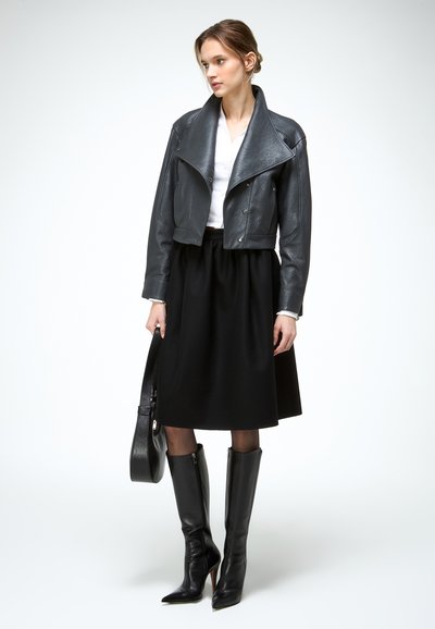 Vrouw gekleed in een zwart leren cropped jack, witte blouse, zwarte rok tot op de knie, zwarte laarzen met hoge hak, een zwarte handtas vasthoudend, staand tegen een witte achtergrond.