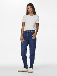Blå denimjeans med rett snitt, kombinert med en enkel hvit bomullst-skjorte og hvite joggesko med brune såler. Enkelt, uformelt antrekk.