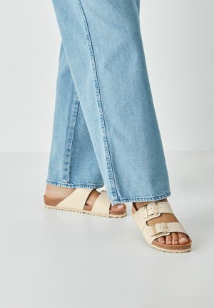 Bézs csúszásgátló szandál két állítható pánttal és ezüst csatokkal, széles, világoskék farmernadrággal párosítva, amelynek nyers szegélyei vannak.