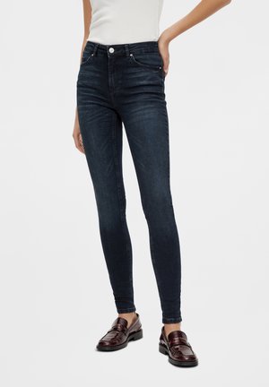 Kobieta ubrana w ciemnoniebieskie obcisłe dżinsy, biały bezrękawnik i ciemnobrązowe skórzane mokasyny, stojąca z jedną ręką na biodrze.