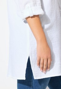 Camicia bianca, strutturata, con maniche arrotolate, spacchi laterali e una vestibilità rilassata, abbinata a jeans scuri, mostrando una mano morbida appoggiata sul tessuto.