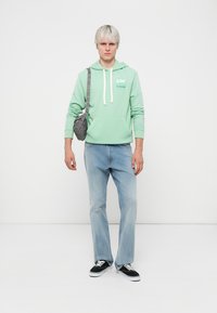 Felpa verde menta con corde bianche e logo, abbinata a jeans azzurri chiari. Il modello tiene una borsa a tracolla grigia e indossa sneakers nere.