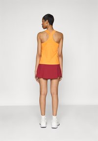 Oranžová športová tielko s prekríženým strihom na chrbte a textúrovanou tkaninou, spárované s priliehavou bordovou sukňou. Biele tenisky dotvárajú outfit.
