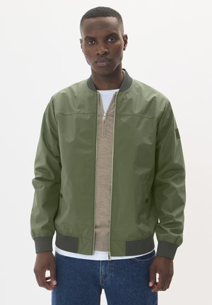 Green bomber jacket with a zip front, knit collar and cuffs, made of smooth material. Paired with a beige sweatshirt and blue jeans.