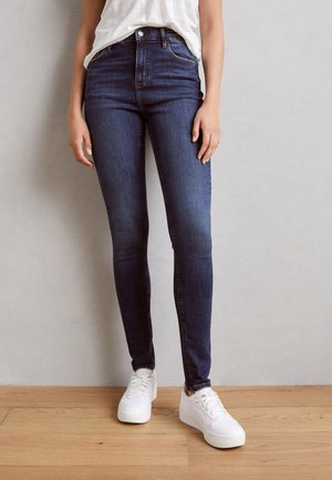 Personne portant un jean skinny bleu foncé, des baskets blanches et une chemise blanche, debout contre un mur gris sur un sol en bois.