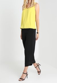 Femme portant un chemisier jaune sans manches, un pantalon large noir, des sandales noires à talons hauts avec des brides, et une montre connectée au poignet gauche.