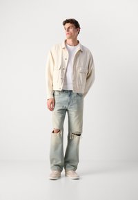 Chaqueta de mezclilla beige claro con dos bolsillos frontales, camiseta blanca, jeans de lavado claro desgastados con grandes rasgaduras en las rodillas, y zapatillas crema.
