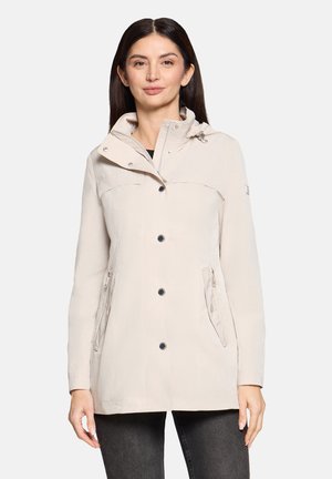 Kobieta o długich ciemnych włosach, ubrana w beżową zapinaną na guziki kurtkę z kieszeniami na zamek i wysokim kołnierzem, stojąca na jasnym tle.