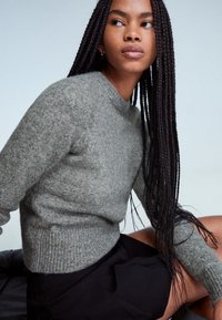 Ung kvinna med långt flätat hår som bär en grå stickad tröja och svart kjol, sitter och tittar åt sidan mot en enkel bakgrund.