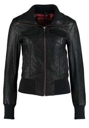 Femme portant une veste noire, un top blanc, un jean bleu foncé avec des revers, et des bottines noires, debout devant un fond uni.