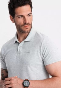 Grå kortärmad polo med knäppt krage och en logotyp på vänster bröst, i kombination med en svart klocka med brun läderrem.