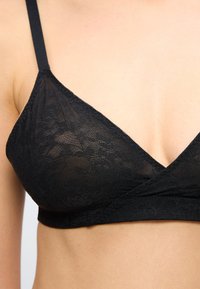 Μαύρο μπραλέτο από δαντέλα με λεπτές τιράντες που φοριέται στον άνω κορμό, δείχνοντας λεπτό διακοσμητικό μοτίβο από λουλουδένια δαντέλα και βαθύ ντεκολτέ σε σχήμα V.