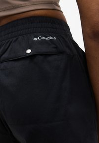 Κοντινό πλάνο ατόμου που φοράει μαύρα σορτς Columbia με ελαστική ζώνη μέσης και πίσω τσέπη ασφαλισμένη με μεταλλικό κουμπί.