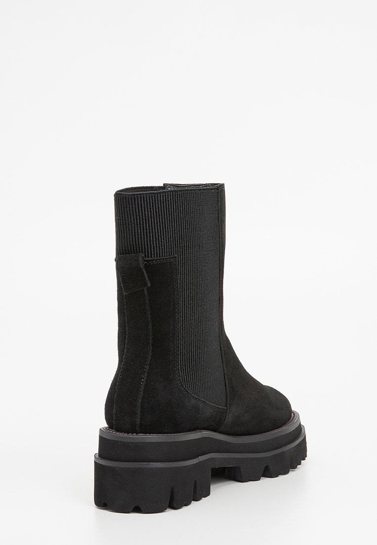 Stivale alla caviglia in suede nero con pannelli laterali elastici a coste e suola spessa in platform. Presenta un tab per facilitare l'indosso e dettagli testurizzati.