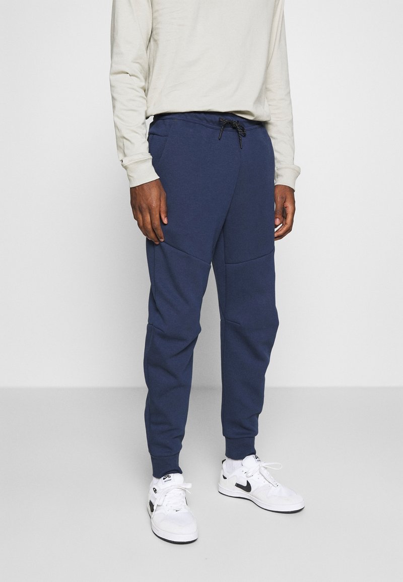 Chandal azul marino hecho de un tejido suave, con cintura ajustable, piernas ceñidas y paneles texturizados a los lados. Zapatillas deportivas blancas.