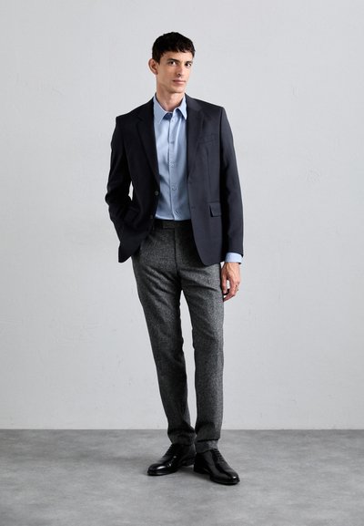 Blazer scuro su misura, camicia azzurra chiara e pantaloni grigi con texture. Scarpe in pelle nera completano l'outfit, su uno sfondo neutro.