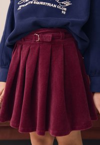 Gonna plissettata in velluto a coste bordeaux con cintura in tessuto e dettaglio fibbia dorata, caratterizzata da una superficie testurizzata e una silhouette svasata.