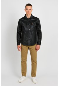 Sort læderjakke med knapper foran, to brystlommer og et mærke med logo. Kombineret med beige bukser og hvide sneakers.