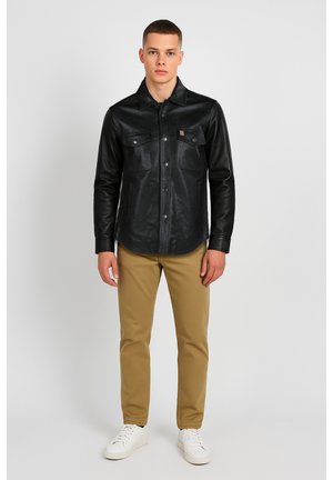 Black leather shirt jacket with a button front, two chest pockets, and a branded patch. Paired with tan pants and white sneakers.