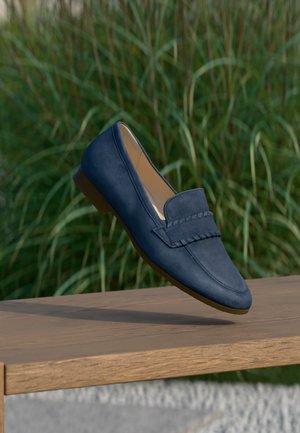 Mocassini blu navy con texture in suede, caratterizzati da una fascia decorativa e un tacco basso, esposti su una superficie in legno con vegetazione sullo sfondo.