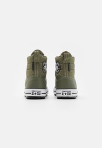 Baskets montantes en toile vert olive avec semelles en caoutchouc, dotées de patchs logo Converse All Star noirs et d'une semelle intermédiaire rayée blanche.