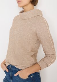 Suéter beige acanalado con cuello alto, con un diseño de punto texturizado y franjas laterales blancas, combinado con jeans de mezclilla azul oscuro.