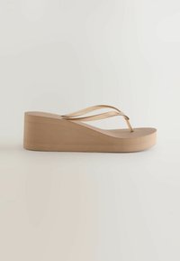 Sandale tong beige à semelle compensée avec des bandes en plastique transparent et une semelle marron, présentée de côté sur un fond uni.