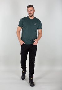 Dunkelgrünes Baumwoll-T-shirt mit Rundhalsausschnitt, kombiniert mit schwarzen Cargo-Hosen mit mehreren Taschen und schwarzen Sneakers mit gelben Akzenten.