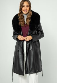 Svart läderjacka med en stor, plyschig svart pälskrage, dubbelknäppta knappar och midja med bälte. Modellen bär en grå polotröja och en vinröd topp.