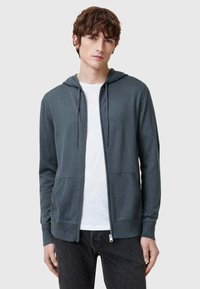 Felpa grigia con zip realizzata in materiale morbido, dotata di zip frontale, due tasche laterali e cappuccio con cordoncino. Indossata sopra una maglietta bianca.