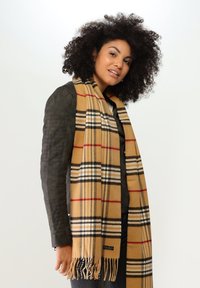 Sciarpa a scacchi beige con linee nere e rosse, bordi frangiati e un'etichetta. Indossata con una giacca scura, mostra una consistenza morbida e una forma rettangolare.