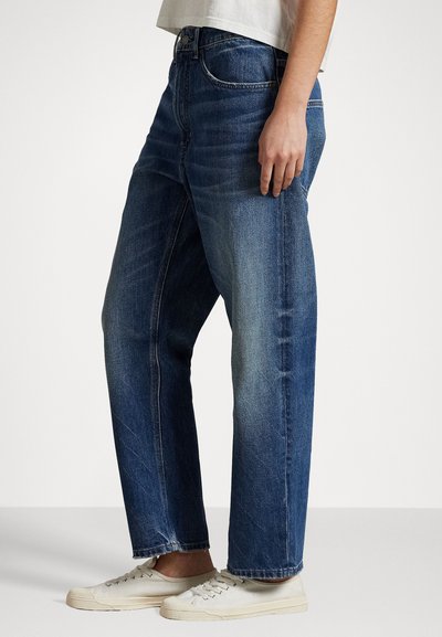 Dark blue denim jeans with a relaxed fit, visible stitching, five-pocket design, and a slightly faded texture at the thighs. Paired with white sneakers.