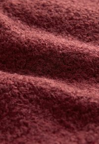 Detailní záběr na měkkou, texturovanou látku s kudrnatými vlákny v hluboké hnědočervené barvě, ukazující jemné záhyby a povrchové detaily.