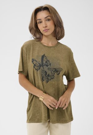 Ung kvinde med skulderlangt hår iført en olivengrøn T-shirt med sorte sommerfugleprint, beige bukser samt guldbøjlearmbånd og øreringe.