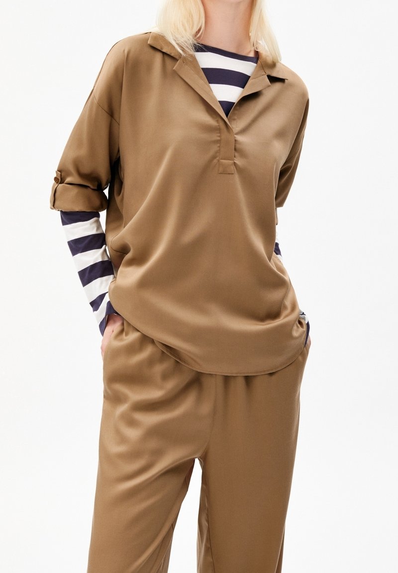 Persona che indossa una camicia a colletto marrone larga e pantaloni con maniche lunghe a righe bianche e nere sotto, con le mani nelle tasche.