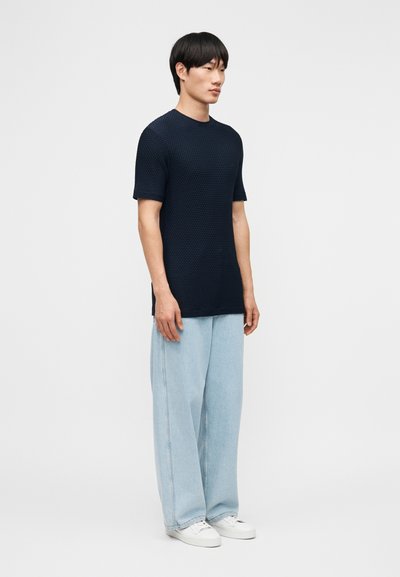 Άνδρας όρθιος, φοράει υφασμάτινο μπλε κοντομάνικο πουκάμισο με υφή, ανοιχτό μπλε τζιν παντελόνι φαρδύ και λευκά αθλητικά παπούτσια, σε απλό φωτεινό φόντο.