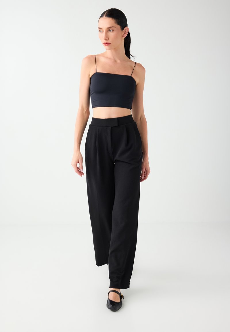 Femme aux cheveux foncés attachés en arrière, portant un crop top noir à fines bretelles, un pantalon noir taille haute et des chaussures noires, marchant vers l’avant.