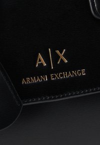 Μαύρη υφή δερμάτινου με χρυσλό λογότυπο A|X και κείμενο "Armani Exchange" κεντημένα στο υλικό.