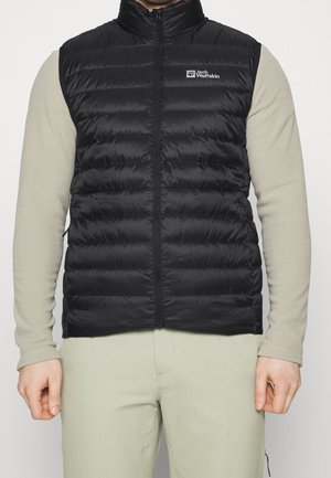 Mann trägt eine schwarze gesteppte ärmellose Weste über einem beigen Langarmshirt und beigen Hosen, steht vor einem einfarbigen weißen Hintergrund.