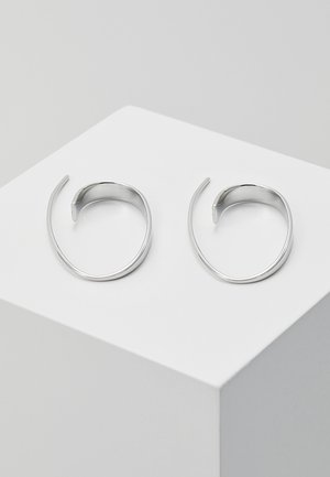 Boucles d'oreilles créoles en argent avec un design ouvert et minimaliste. Présentent des formes lisses et courbées ainsi qu'une texture uniforme, reflétant la lumière de manière subtile.
