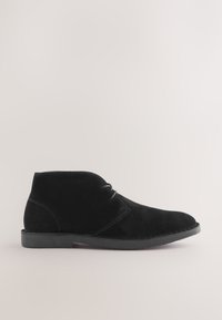 Bottine basse noire en daim avec deux œillets pour lacets, semelle cousue et petit talon sur un fond clair uni.