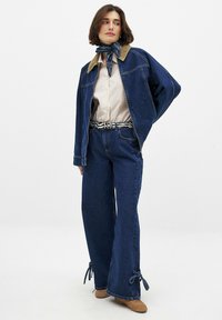 Denim jakna s kordno ovratnikom, prugasta srajca, široke denim hlače z vezalkami na robu in modra bandana okoli vratu.