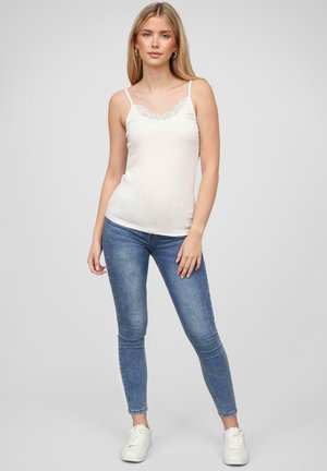 Weißes Trägertop mit Spitzenbesatz, dünnen Trägern, kombiniert mit blauen Skinny-Jeans und weißen Sneakers. Glattes Material mit tailliertem Schnitt.