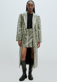 Manteau et jupe à motif serpent dans des tons de vert et d'argent ; présente une silhouette longue, des revers bien marqués et une fente sur le côté ; associé à des bottes noires montantes.