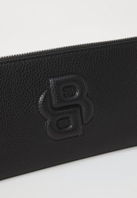 Black leather wallet with textured finish, featuring a raised logo detail. Zipper closure along the top edge. Simple, functional design.